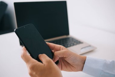 Klas gömlekli tanınmayan yönetici cep telefonunda hafif iş yerinde dizüstü bilgisayarla masada otururken kısa mesaj atıyor