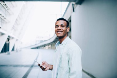 Beyaz tişörtlü ve tişörtlü, şehir caddesinde binaların yanında tek başına dururken akıllı telefon kullanan pozitif siyah adamın yan görüntüsü.