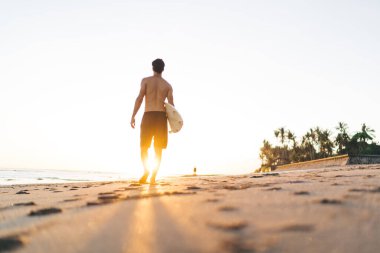 Alçak açılı, formda, şortlu, isimsiz bir erkek sporcunun kumlu sahilde sörf tahtası ve okyanus kenarında ayak izleriyle dolaşması.