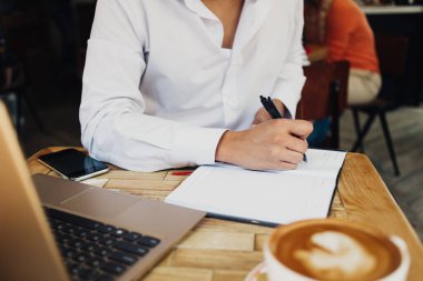 Beyaz tişörtlü, laptoplu, kahveli ve not defterine notlar yazan isimsiz bir öğrenci. 