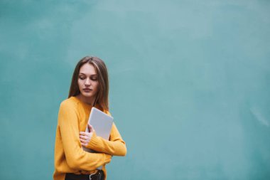 Uzun siyah saçlı, parlak kazaklı genç bayan elinde dizüstü bilgisayarla turkuaz duvarın yanında duruyor.