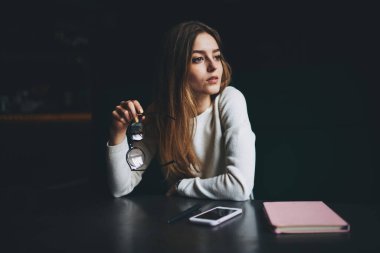 Ciddi bir kadın, gündelik kıyafetler içinde, uzun saçlı, karanlık bir masada oturuyor, elinde gözlük, elinde telefon ve gözlüklerle dinleniyor.