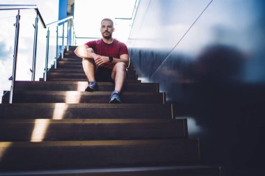 Spor giysiler ve spor ayakkabılar giyen genç sakallı bir erkeğin merdivenlerde oturup ciddi bir yüzle kameraya bakmasından.