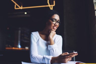 Siyah saçlı, pozitif Afrikalı bir kadın. Gündüz vakti masa başında oturmuş, akıllı telefonlara bakıyor.