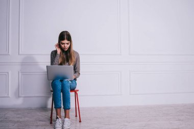 Düşünceli kadın uzaktan kumandalı, gündelik kıyafetli, sandalyeye oturmuş saçına dokunuyor ve dizüstü bilgisayarla yeni bir proje üzerinde çalışıyor.