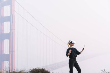 San Francisco 'da köprünün yakınındaki tepede dururken, modern bir aletle sanal gerçekliği deneyimleyen kimliği belirsiz bir kadının yan görüntüsü.