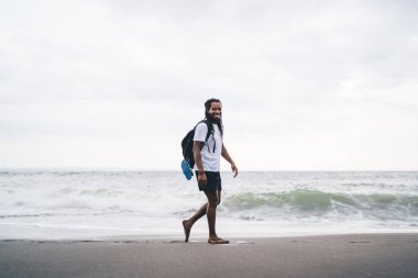 Sırt çantalı, sırtüstü gülümseyen ve deniz kıyısında kameraya bakan Afro-Amerikan erkek.