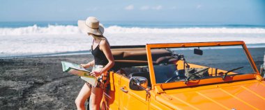 Sıradan giyinmiş, şapkalı, elinde harita olan, deniz kıyısında arabanın yanında duran ve denize bakan tanınmamış genç bir bayan turist.