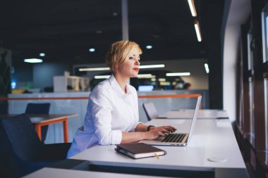 Konsantre olmuş orta yaşlı, sarışın kadın çalışan resmi giysiler içinde masaya oturmuş dizüstü bilgisayarla yazı yazarken gözlerini kaçırıyor.