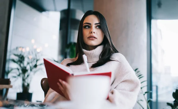 Low angle of pensive woman wearing warm clothes thinking on new article while sitting at table with cup of hot drink