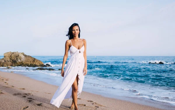 Portrait of Latin woman dressed in comfortable white sundress walking at coastline beach during pastime near Caribbean sea
