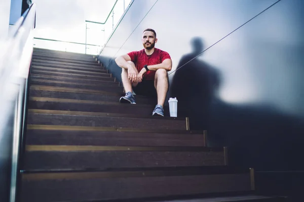 From below of male athlete in sports outfit and sneakers sitting on steps and looking away while relaxing after fitness class