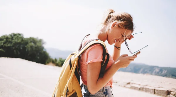 Side view of cheerful female tourist with backpack browsing smartphone and standing against mountains and plants under cloudless sky in sunny day