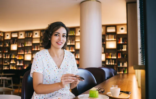 Kitapçıda kameraya bakarken masada oturan tatlı kremalı kek ve bir fincan kahveyle samimi bir bayan.