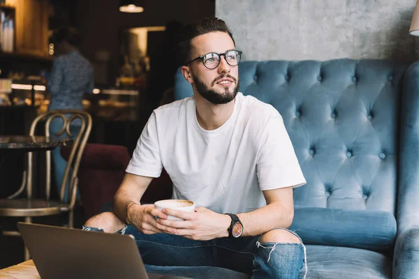 Thoughtful male programmer with cappuccino cup in hands thinking about project idea during remote job in coworking space