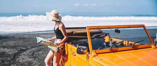 Sıradan giyinmiş, şapkalı, elinde harita olan, deniz kıyısında arabanın yanında duran ve denize bakan tanınmamış genç bir bayan turist.