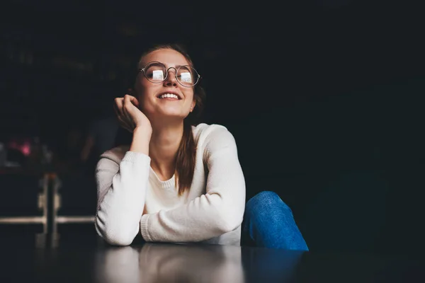 低い角度の若いです幸せな女性でホワイトジャンパーと眼鏡で座ってテーブルと暗い部屋のカメラを見て — ストック写真