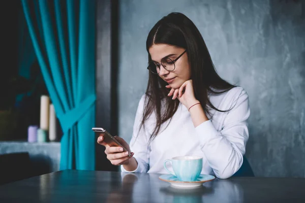Pensive female in casual outfit with eyeglasses sitting at table with cup of tea leaning on hand and browsing mobile phone