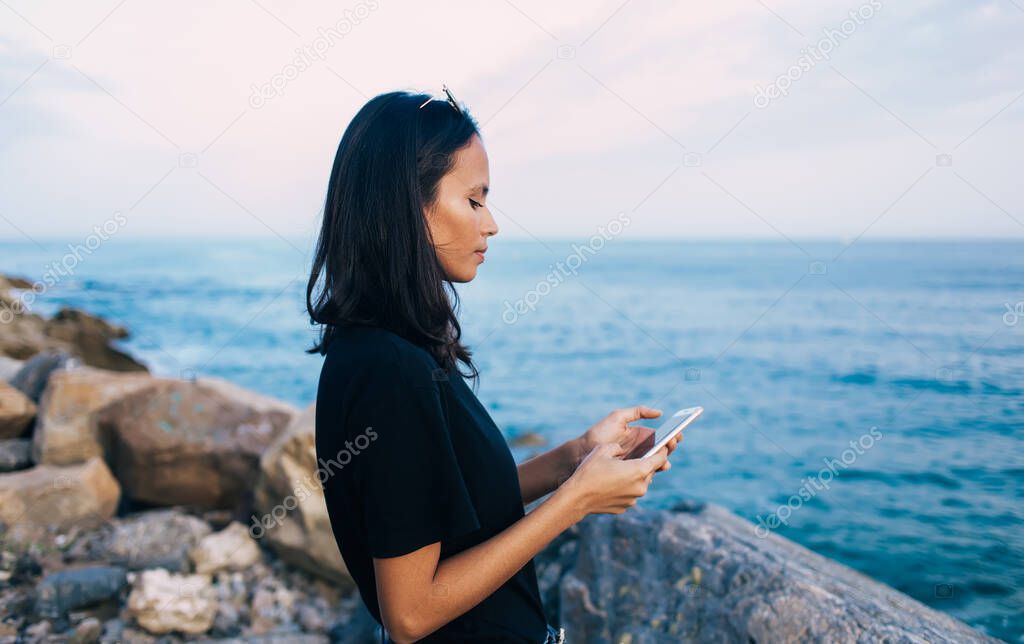 Side view of attractive female millennial using smartphone device for checking coastline location during summer vacations