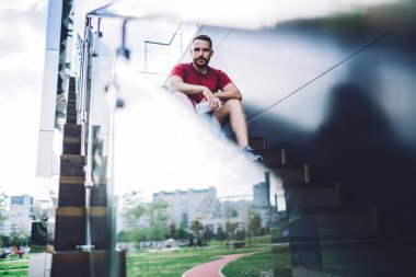 Spor giyim kuşamında elinde plastik su şişesiyle merdivenlerde oturmuş kameraya bakan ciddi bir erkek koşucunun camından.