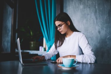Hasretli, uzaktan kumandalı, gözlüklü bir kadın bir fincan kahveyle masada oturuyor ve iş sırasında laptoptan bilgi arıyor.