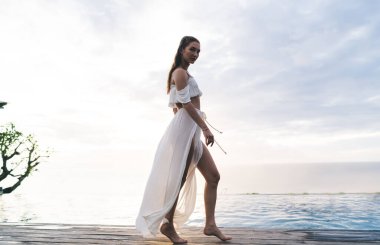 Beyaz elbiseli, çıplak ayaklı, patikada dikilirken kameraya bakan zarif kadının alçak açılı yan görüntüsü.