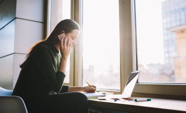 Sıradan kıyafetli, pencerenin yanında dizüstü bilgisayarla oturan ve cep telefonuyla konuşurken bilgi yazan serbest çalışan kadın görüntüsü