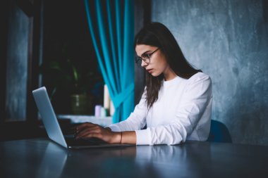 Genç odaklı serbest çalışan, gündelik giysiler içinde, gözlüklü, masada oturan ve modern çalışma alanında çalışırken dizüstü bilgisayarını karıştıran bir kadın.