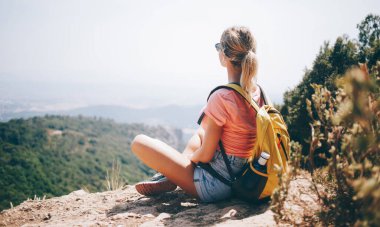 Sırt çantalı, yaz kıyafetleri ve güneş gözlükleriyle tepede otururken resimli manzaranın tadını çıkaran isimsiz genç bir gezginin arka planı.