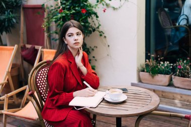 Süslü giysiler içindeki düşünceli genç bayan bir fincan kahveyle masada otururken ve terasa bakarken deftere yazı yazıyor.