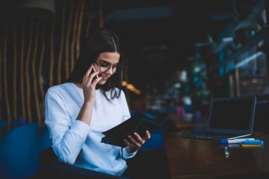 Neşeli, uzaktan kumandalı, günlük kıyafetli genç bir kadın. Çalışırken cep telefonuyla konuşuyor ve not defterindeki notları okuyor.
