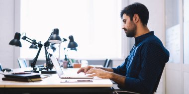 Genç düşünceli erkek yöneticinin dizüstü bilgisayarda oturup not defterleri ve modern ofis lambalarıyla yazı yazışının yan görüntüsü.