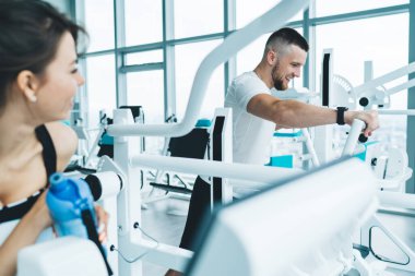Spor kıyafetleri içinde gülümseyen sporcunun eliptik ekipman üzerinde kardiyo egzersizi yaparken spor salonunda cam duvarlı kişisel antrenörle antrenman yaparken yan görüntüsü.