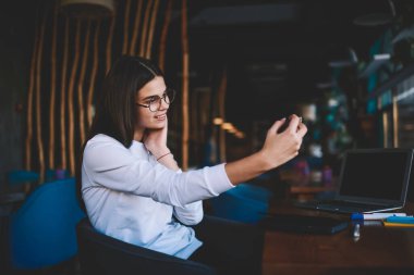 Beyaz kazaklı, gözlüklü, cep telefonlu ve kafeteryada dizüstü bilgisayarla selfie çeken genç bir kadının yan görüntüsü.
