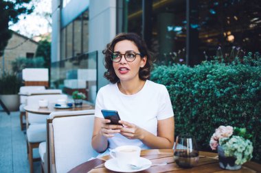 Beyaz gömlekli, şık gözlüklü, cep telefonlu ve açık hava restoranında otururken yüzünü çeviren çekici bir kadın.