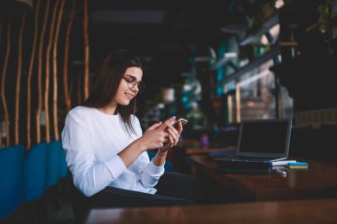 Genç iyimser kadın serbest çalışan, günlük kıyafetler giyen, gözlüklü, dizüstü bilgisayarlı ve cep telefonu kullanan.