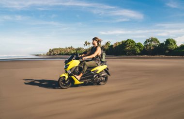 Güneşli bir günde dalgalı okyanusun yanındaki muhteşem kumlu sahilde hızlı motosiklet süren bronzlaşmış genç erkek gezginin yan görüntüsü.