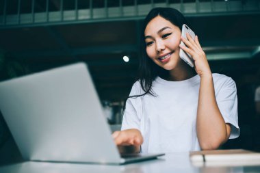 Neşeli etnik kadın serbest çalışan, dişlek gülüşlü, iş sırasında cep telefonuyla konuşurken modern dizüstü bilgisayarlara göz atan.