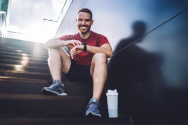 Aktif giyimli neşeli erkek koşucunun plastik şişeyle merdivenlerde oturması ve antrenmandan sonra kameraya bakması.