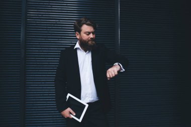 Siyah takım elbiseli ciddi sakallı bir erkek. Kol saati saatine bakıyor. Siyah duvarın yanında dikilirken saati kontrol ediyor.