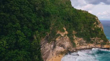 Yeşil ağaçlarla kaplı dağın insansız hava aracının muhteşem manzarası ve bulutlu bir günde dalgalanan dalgalarla okyanusun kumlu kıyıları.