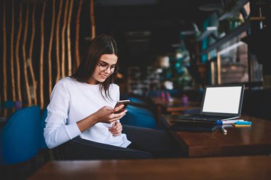 Gülümseyen kadın uzaktan kumandalı günlük giysiler içinde gözlüklü iş sırasında dizüstü bilgisayarla masada oturuyor ve cep telefonuyla sörf yapıyor.