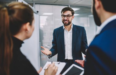 İsimsiz iş ortakları pano ve tabletle ayakta dururken aynı zamanda ofisteki resmi kıyafetler içindeki neşeli iş adamlarından yeni proje fikirlerini dinliyorlar.