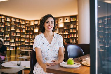 Süslü giyimli genç bir bayan masada lezzetli kremalı pasta ve kafede bir fincan kahveyle kameraya bakıyor.