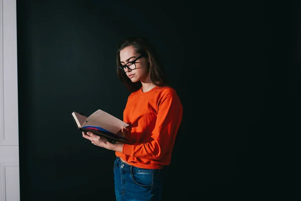 Focused dark haired female in casual wear and spectacles attentively flipping planner while spending time in studio on black background