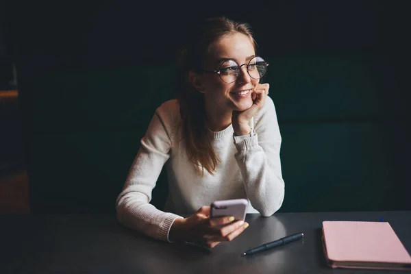 Positive female in casual clothes with eyeglasses looking away sitting at table with notepad leaning on hand using mobile phone during rest