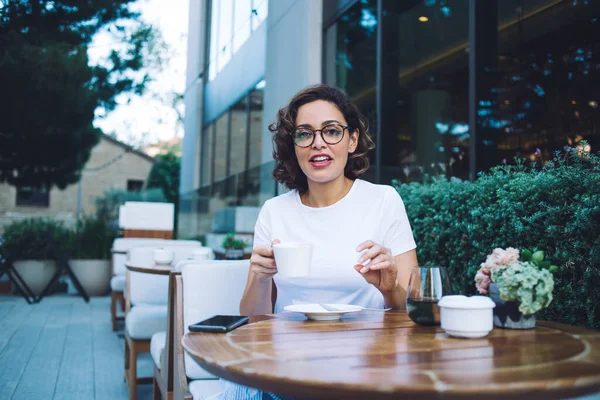 Beyaz gömlekli mutlu bir bayan aromatik taze kahve içiyor ve modern açık hava kafesinde mola verirken kameraya bakıyor.