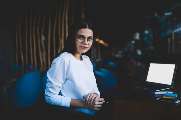 Relaxed brown haired lady in casual wear and eyeglasses looking at camera while browsing laptop for remote work in restaurant