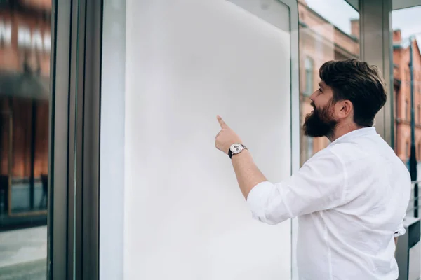 Side view of smiling bearded man standing on bus stop and pointing finger on whiteboard in city street on blurred background