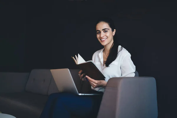 Positive ethnic female in formal outfit smiling and looking at camera while reading book and working on project on laptop